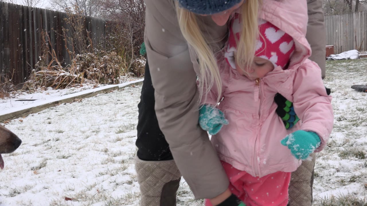 Adult woman steadies a toddler in a snowy yard.