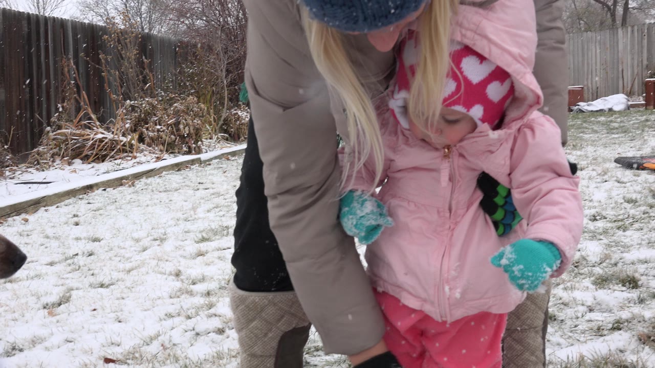 The toddler looks down at her mittened hands while the adult supports her from behind in the snowy Westminster yard.