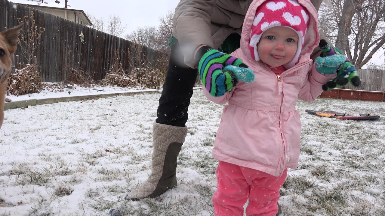 Toddler in pink winter clothing stands in snowy yard with adult hands nearby.