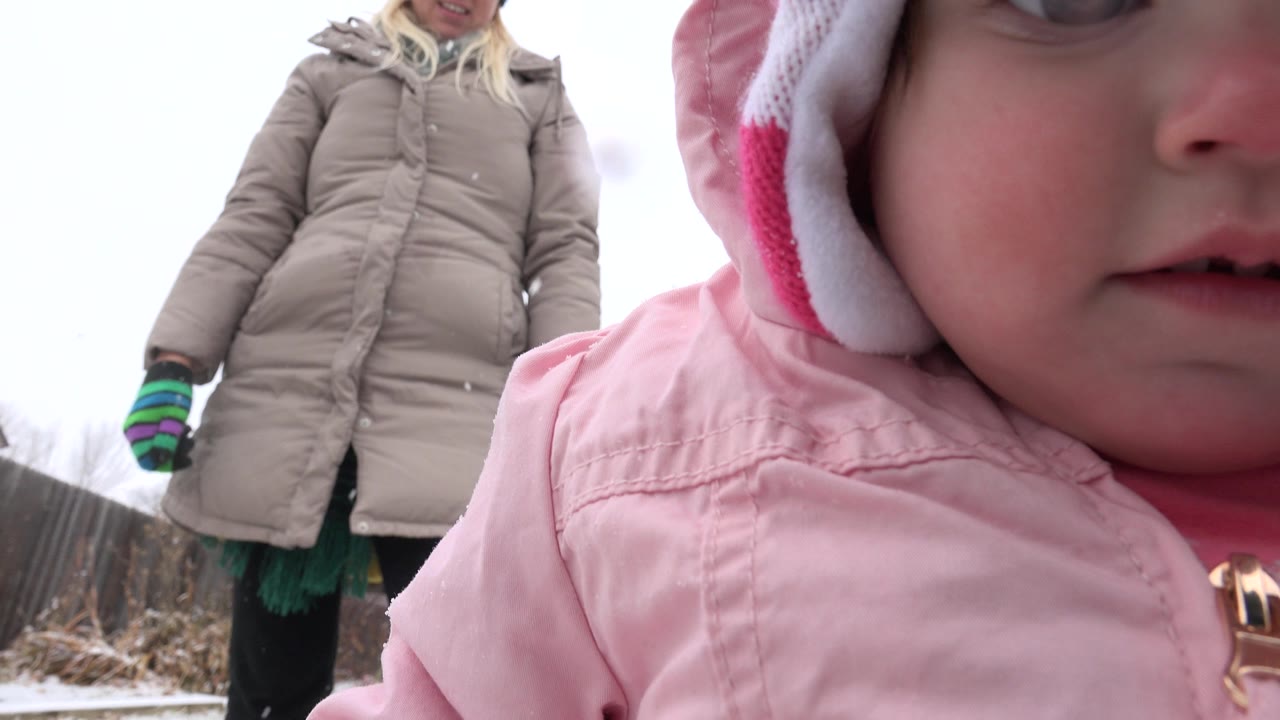 Toddler in a pink winter coat close to the camera with an older woman standing behind.