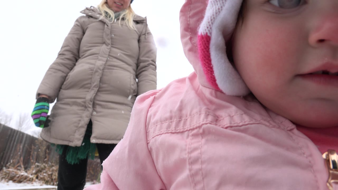 At 0:01 in Westminster, Colorado, the toddler remains very close to the lens with only part of the face visible, while the older woman stands in the background wearing a beige coat and striped gloves.