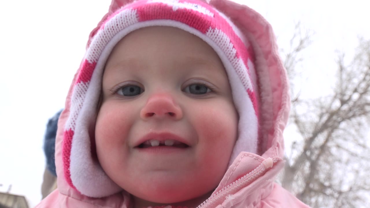 A smiling baby girl in a pink coat and striped winter hat looks toward the low camera in Westminster, Colorado, with bare trees and pale sky behind her.
