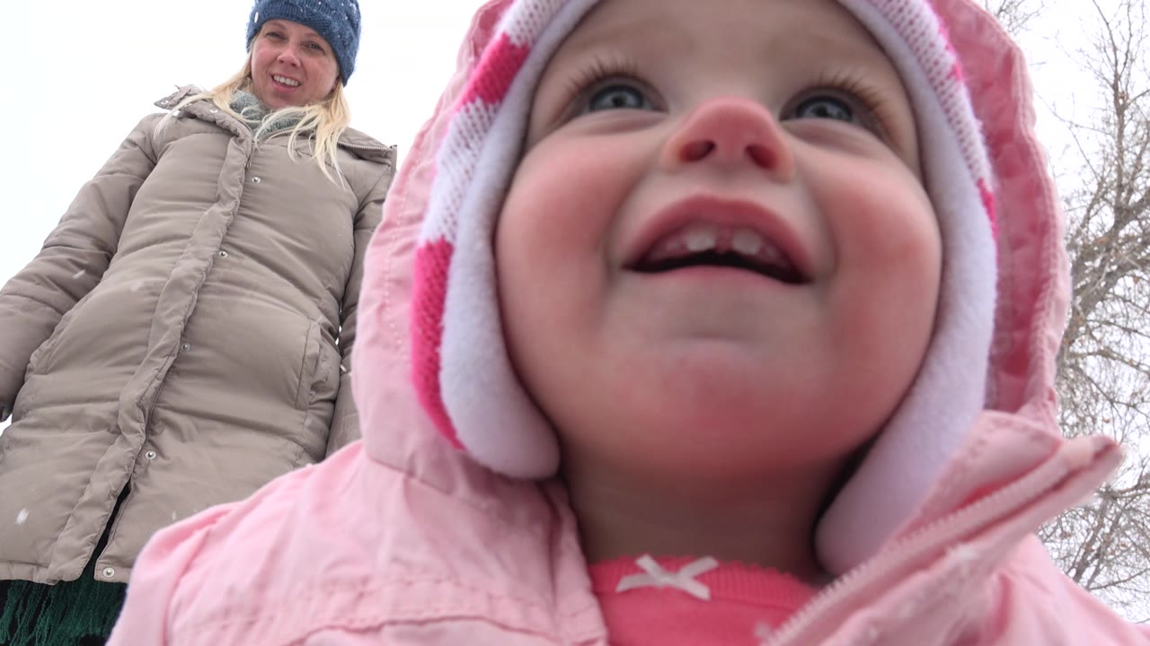 Toddler in pink winter clothing smiles close to the camera with a woman behind her.