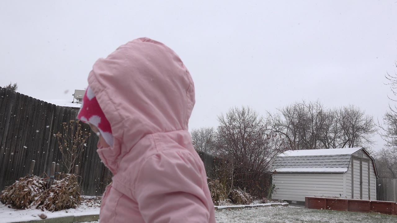 Child in pink snowsuit shown in profile beside a snowy fenced yard.