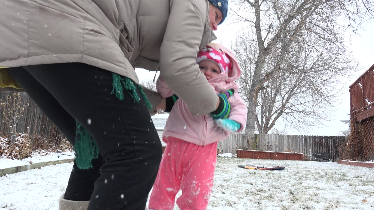 Woman in tan coat bends toward baby in pink in snowy backyard.