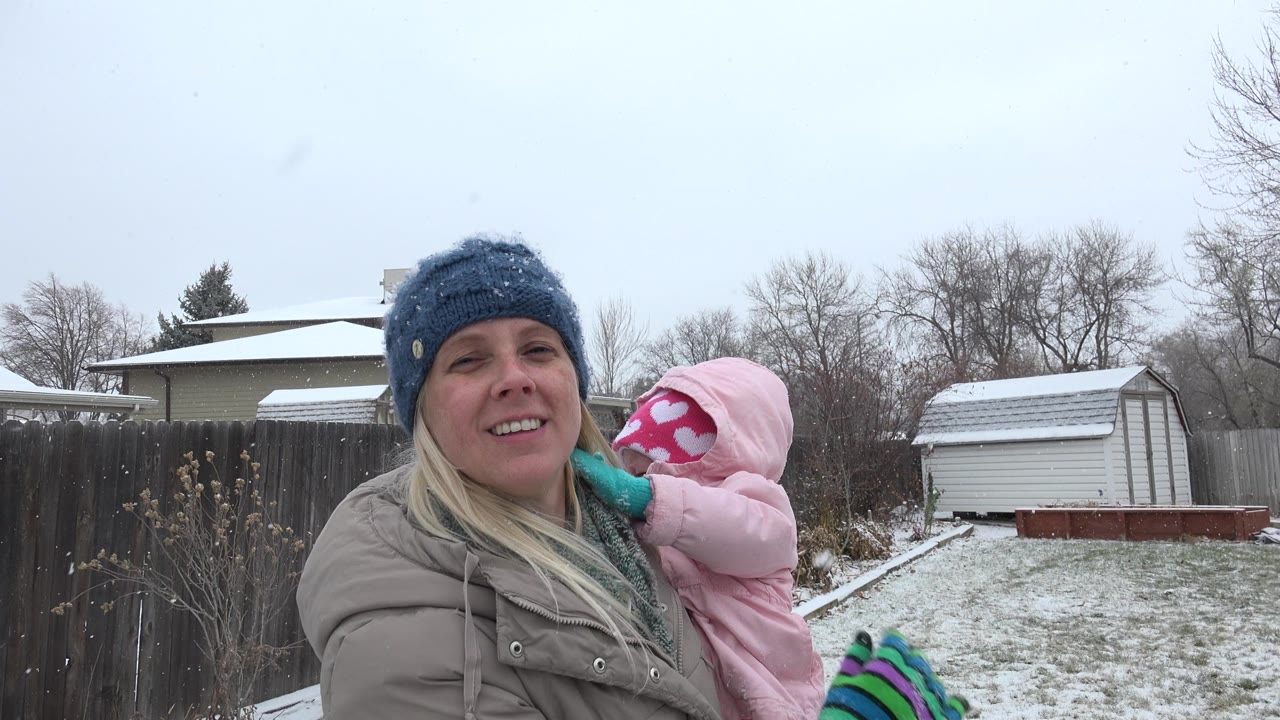 Woman holding a child in pink winter clothing during snowfall in a backyard.
