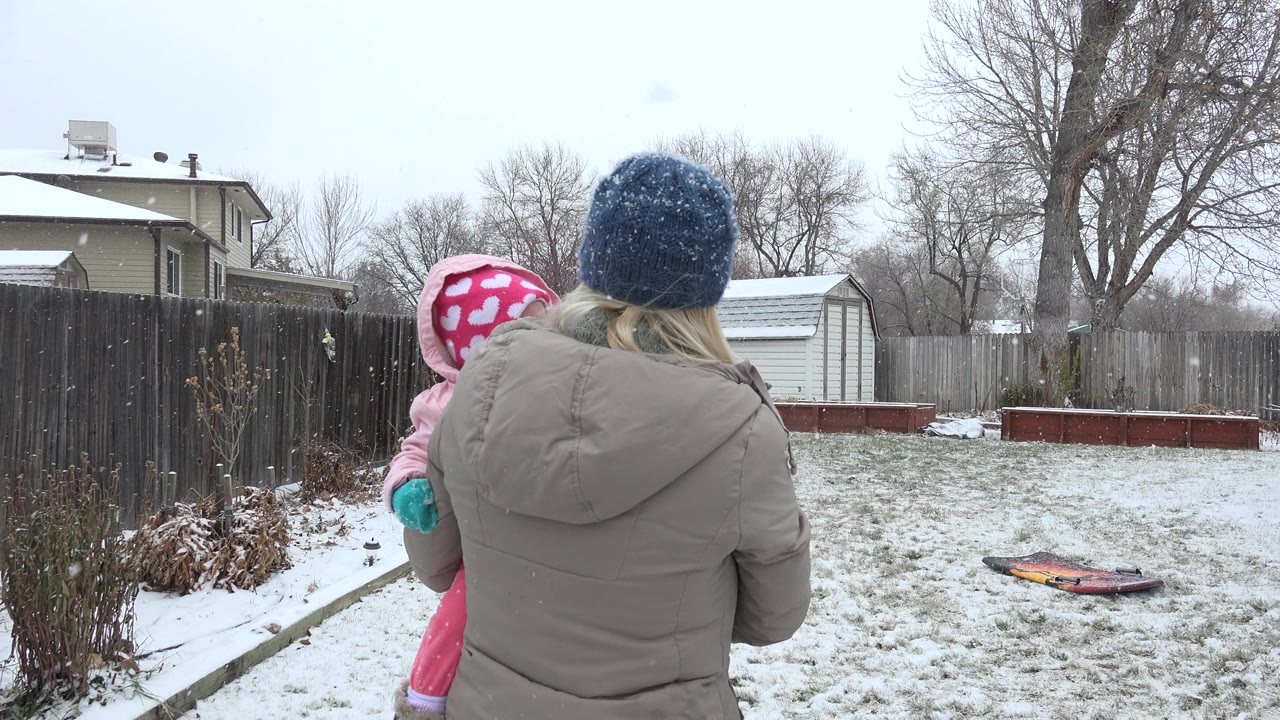 In Westminster, Colorado, a woman in a gray coat holds a child in pink winter clothes near the left side of a lightly snow-covered fenced backyard.