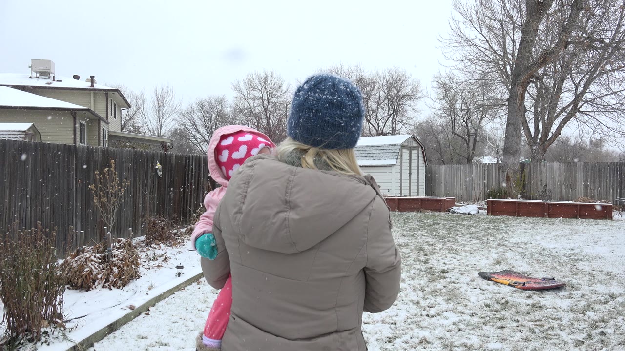 In Westminster, Colorado, the mother and child remain near the foreground as light snow falls over a fenced yard with a white shed and raised beds in the background.