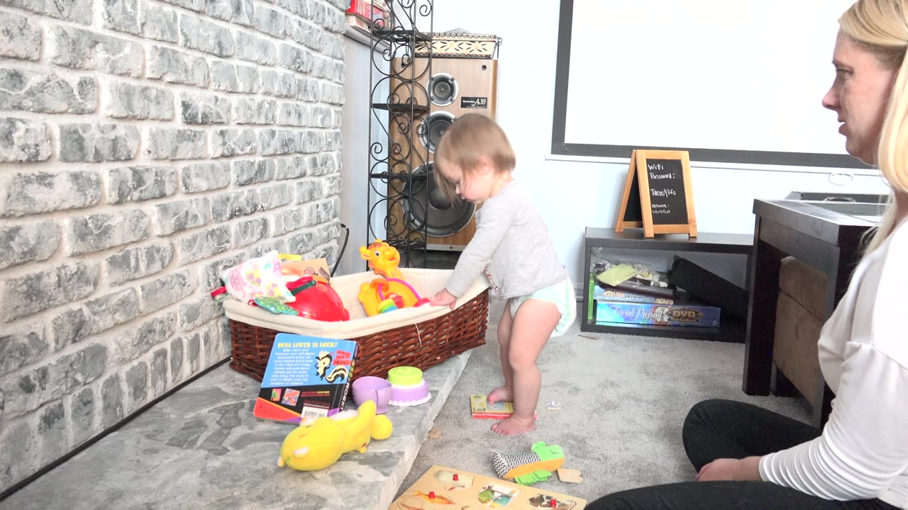 In Westminster, Colorado, a toddler stands at a wicker toy basket as a seated blonde adult is cropped along the right side of the bright home interior frame.