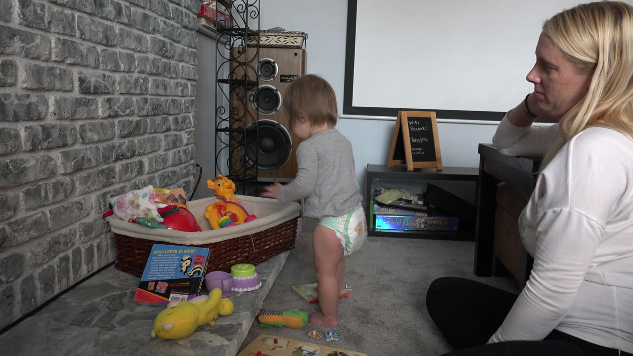 Woman in white shirt watches toddler beside toy bin.