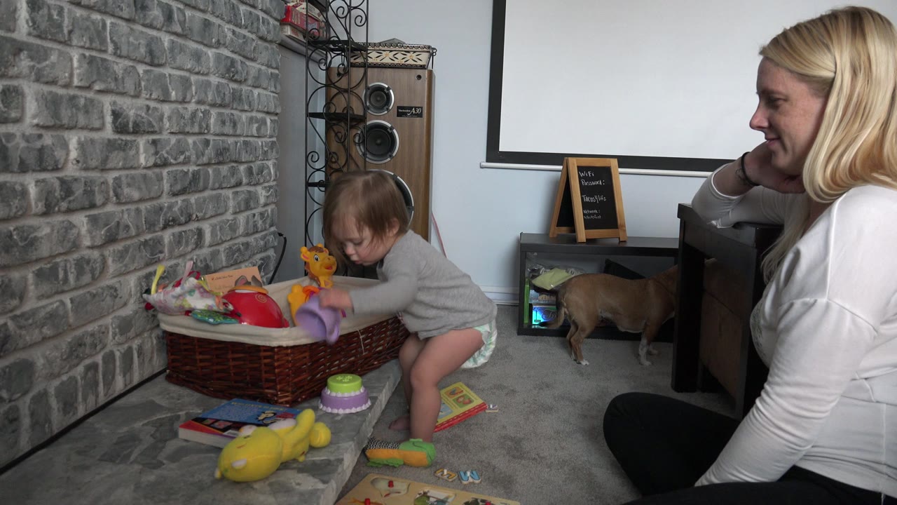 Baby stands beside a basket of toys while a mother watches from the side.