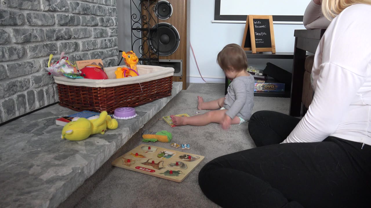 Baby seated on carpet near toys and a partially visible adult.