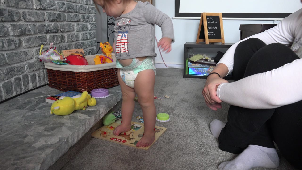 The Westminster home video frame shows the toddler standing on a small board near toy pieces, with the adult seated on the carpet and cropped at the side.
