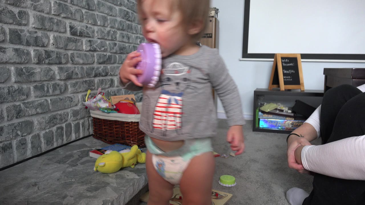 Toddler holding a purple toy in a living room while a woman sits on the floor nearby.
