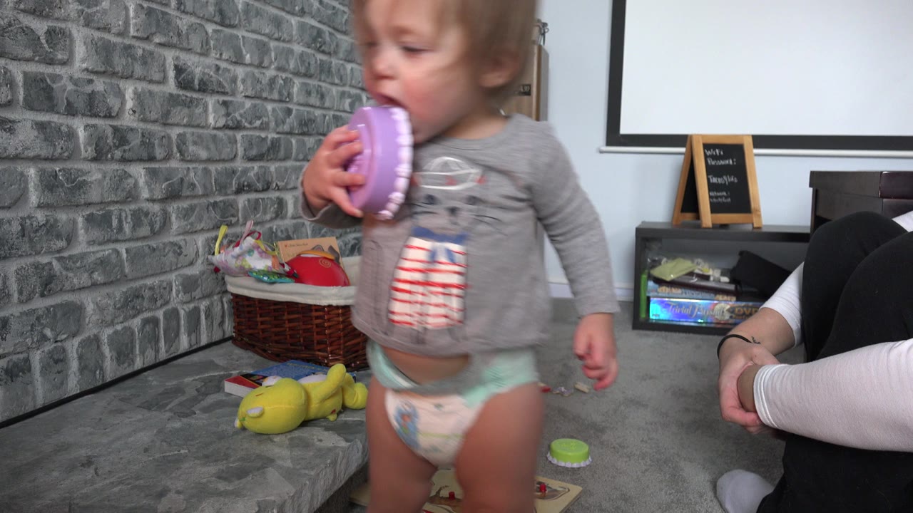 The toddler stands centered in the Westminster, Colorado home interior with the purple toy at their mouth, while the seated woman is partly hidden behind the child.