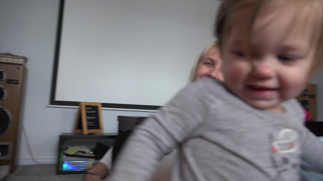 Baby girl close to camera with smiling woman behind her in a bedroom.