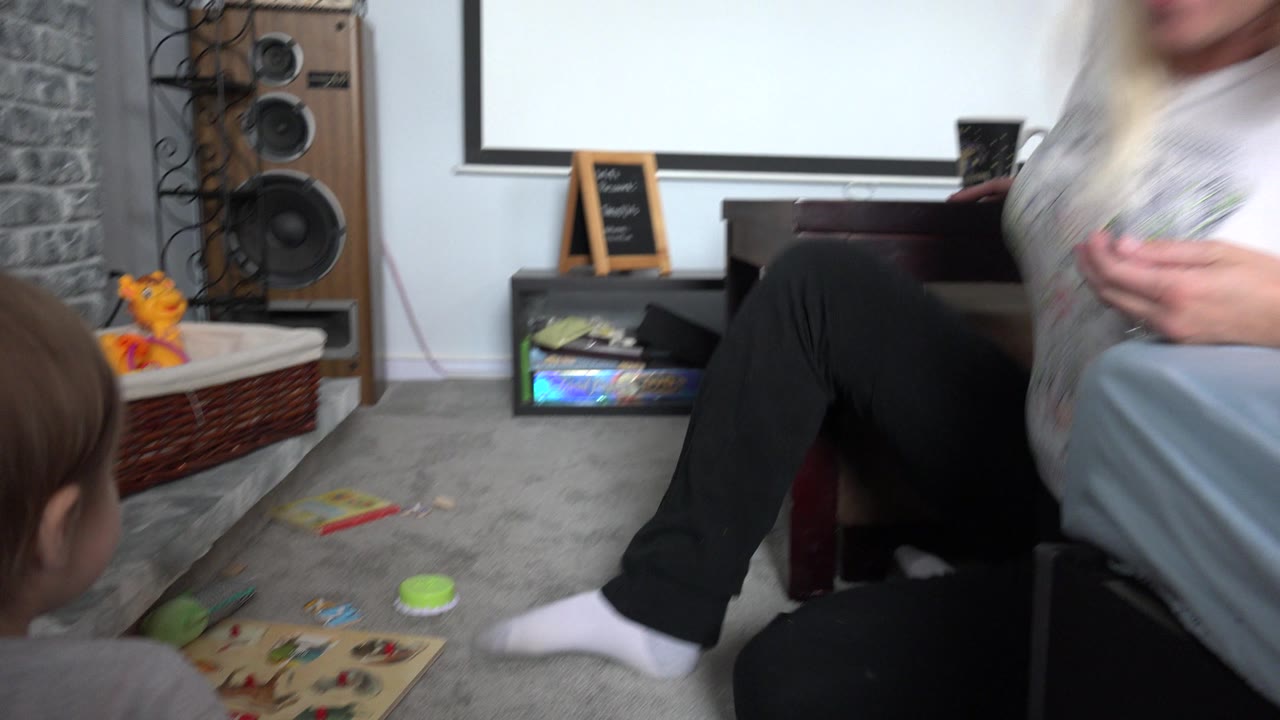 The Westminster, Colorado home movie frame shows the toddler facing an adult near a wooden toy board, with stuffed toys, a basket, speakers, and a stone hearth in the room.