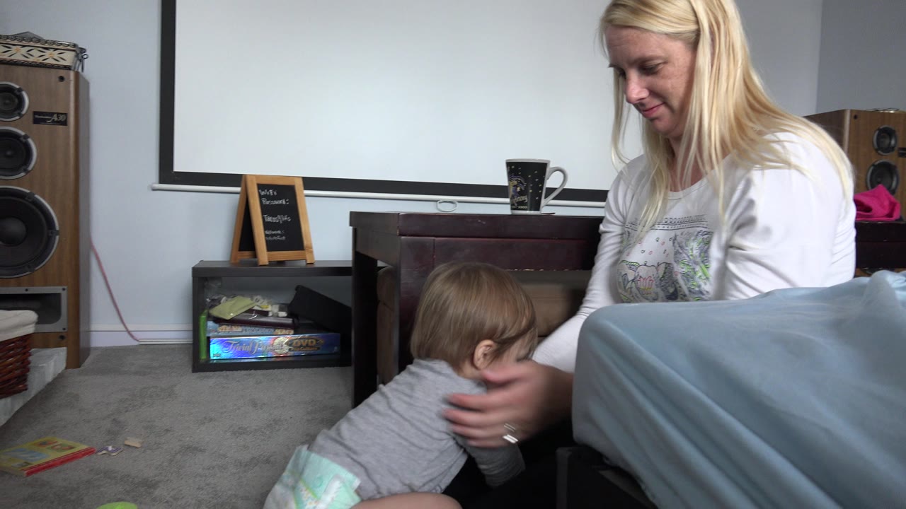 Blonde woman reaches toward a baby on a carpeted living room floor.