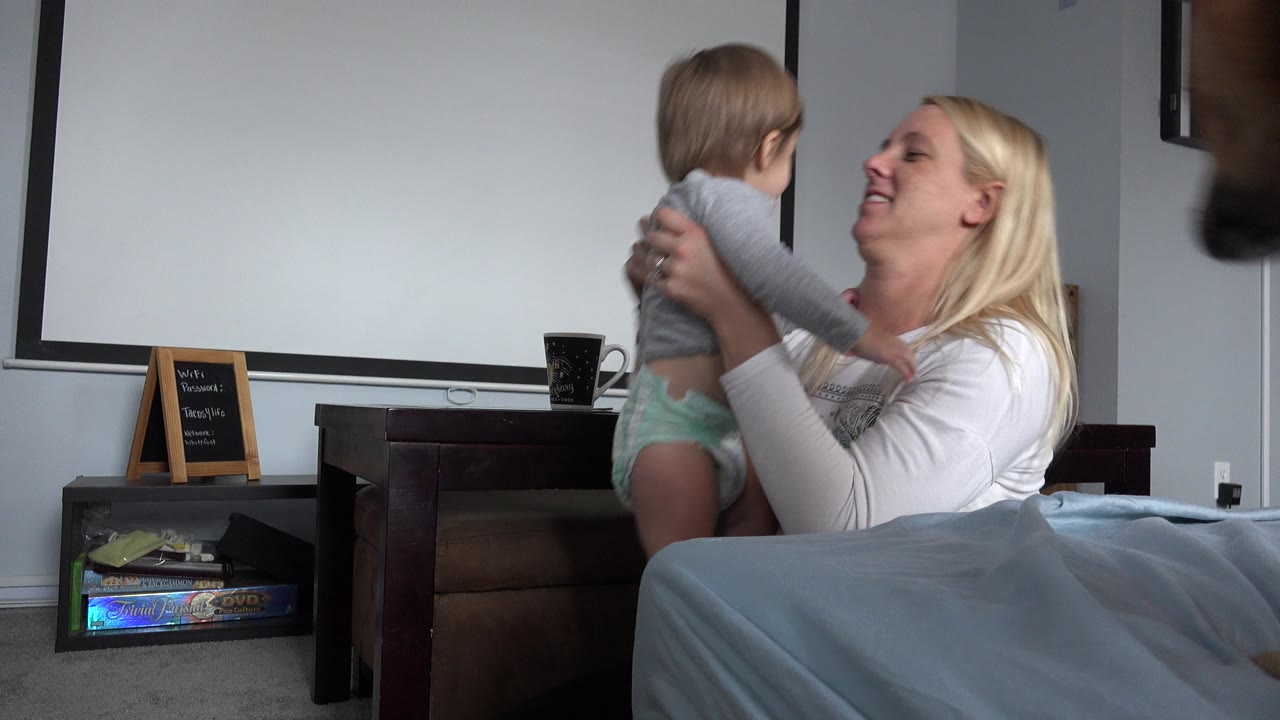 In Westminster, Colorado, a blonde mother holds a baby facing her beside a bed, with a projection screen, shelf, and small chalkboard behind them.