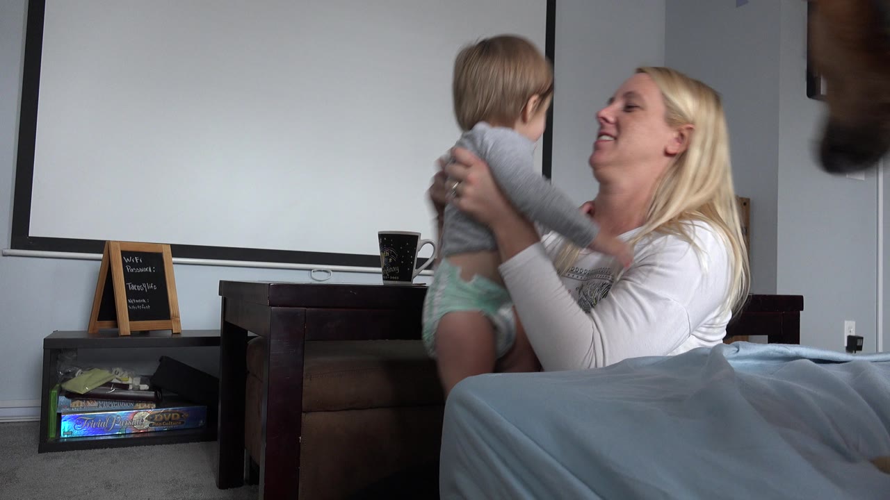 The mother smiles toward the baby in the Westminster home interior as part of a brown dog appears close to the camera at the right edge.