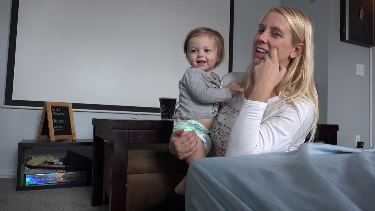 Mother holding a baby beside a bed in a bedroom.