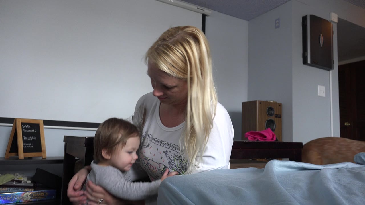 Mother seated beside a toddler near bedding in a home interior.