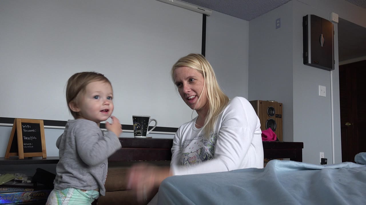 Toddler standing beside a woman on a bed in a home interior.