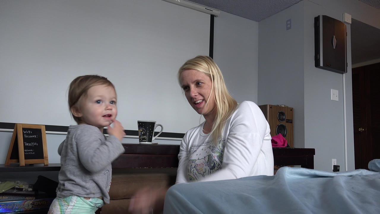 In Westminster, Colorado, the toddler faces forward with one hand near the mouth while the woman in a white shirt looks toward the child from the bed.
