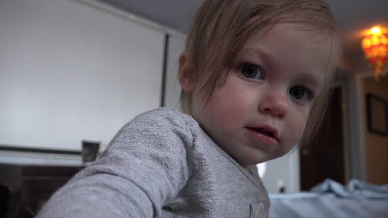 The toddler shifts to the right side of the frame and reaches near the camera, with a warm ceiling light and home furnishings visible behind her in Westminster, Colorado.