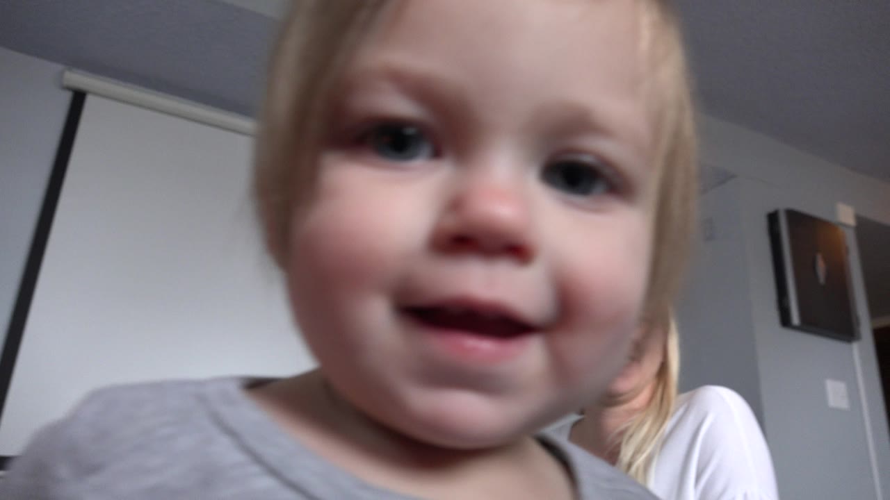 A blonde toddler in a gray shirt looks into a low indoor camera in Westminster, Colorado, while a blonde woman is partly visible behind her near white walls and window shades.