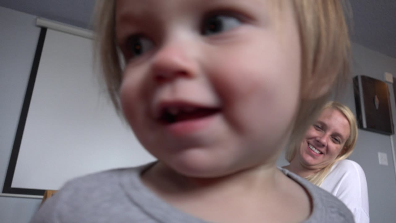 The toddler turns toward the right edge of the frame, revealing more of the smiling blonde woman seated in the Westminster, Colorado room behind her.