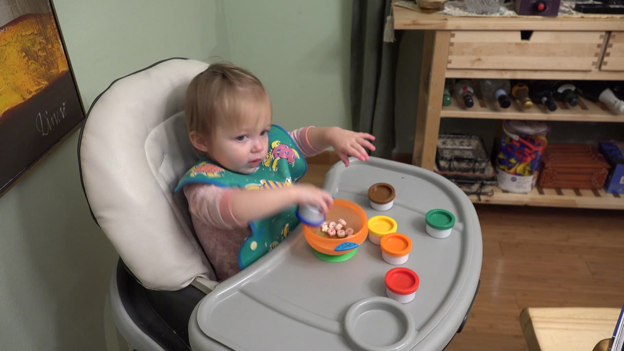 The child remains seated in the Westminster home interior while holding a blue toy near an orange bowl on the high chair tray.