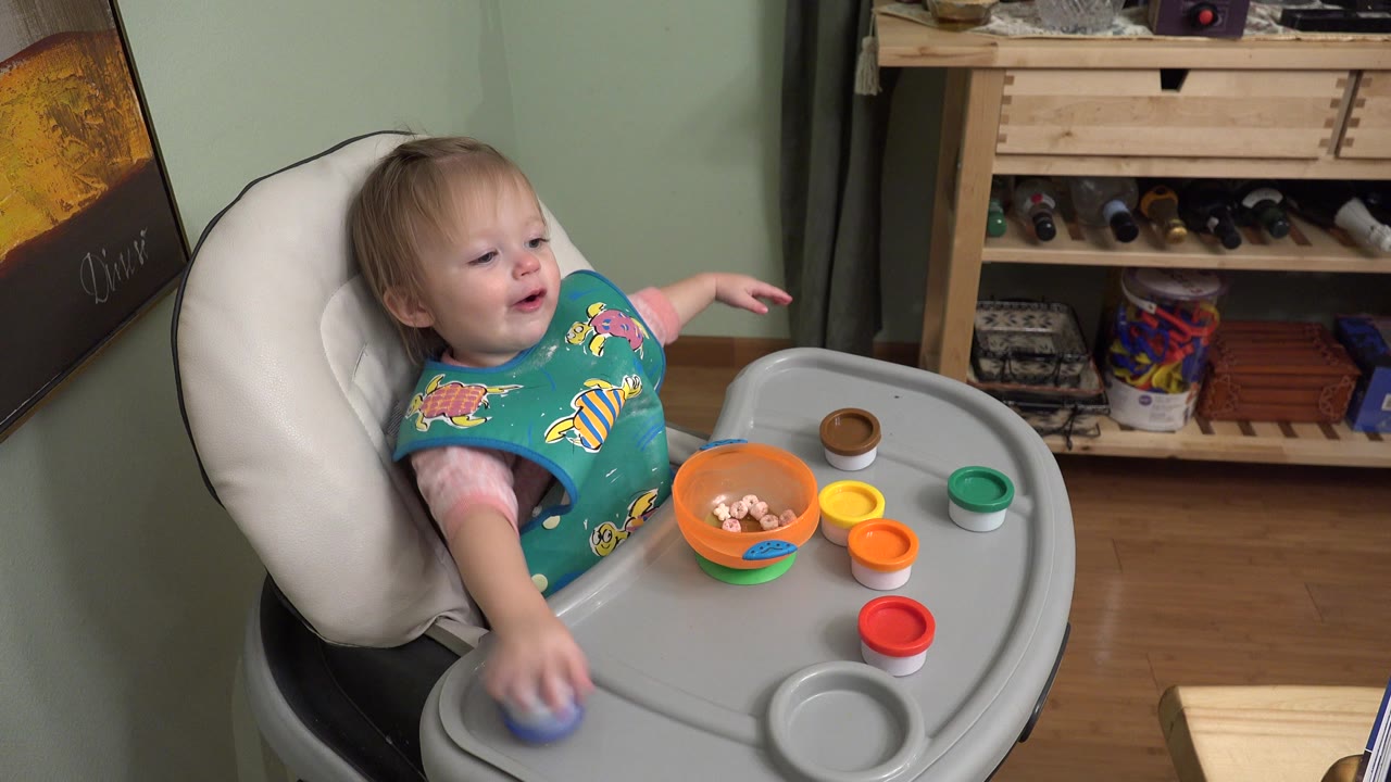 At 0:00 in Westminster, Colorado, a young child in a teal bib sits in a high chair and reaches across a gray tray with an orange bowl and colorful lids.