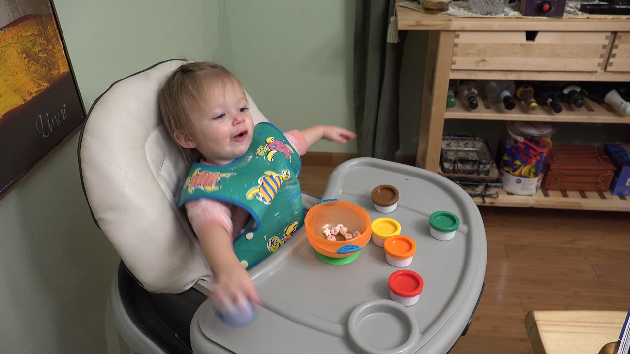 At 0:01 in Westminster, Colorado, the child leans back in the high chair while holding a small blue object near the front edge of the tray.