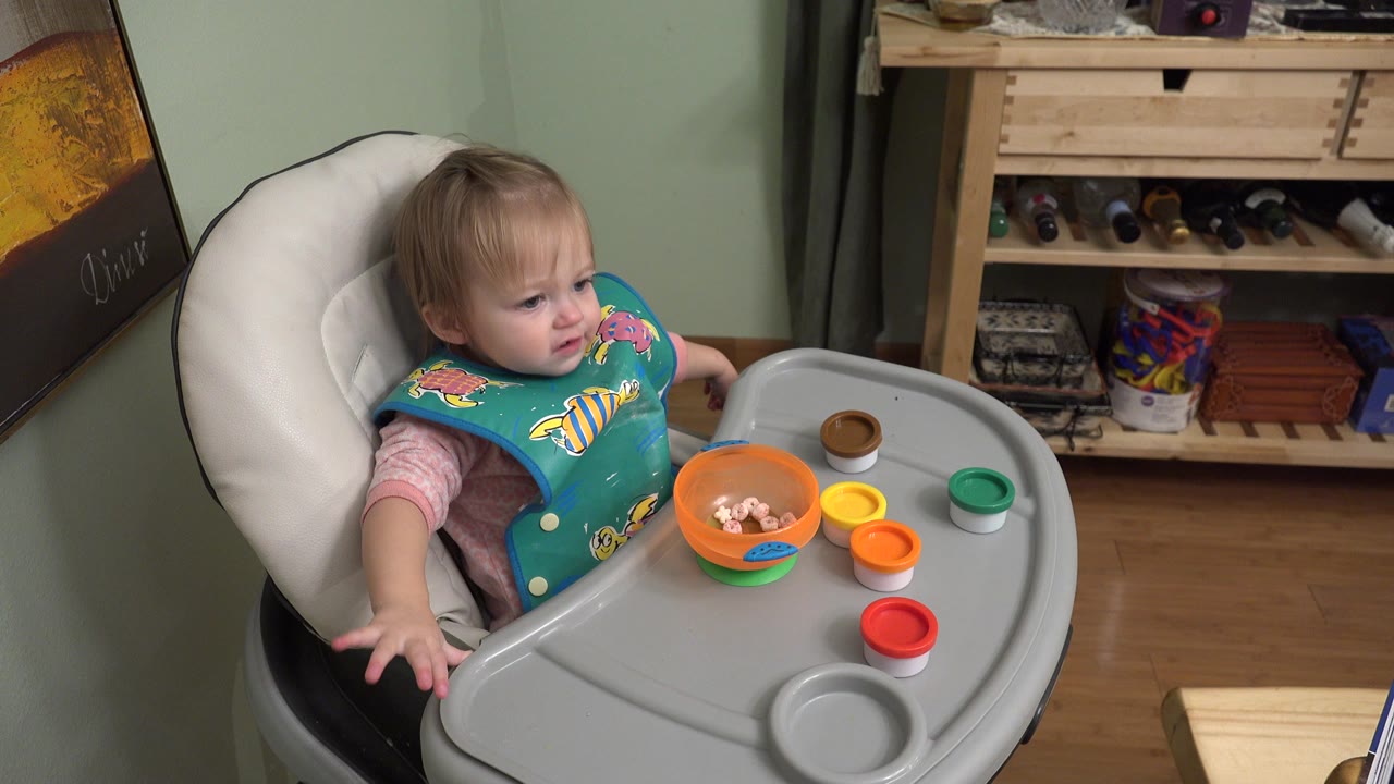 Blond toddler in a high chair with an orange bowl and colorful cups on the tray.