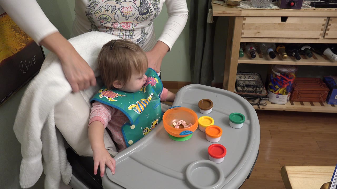 A baby sits in a gray high chair in a Westminster, Colorado home while an adult woman in a white top leans close behind the chair.