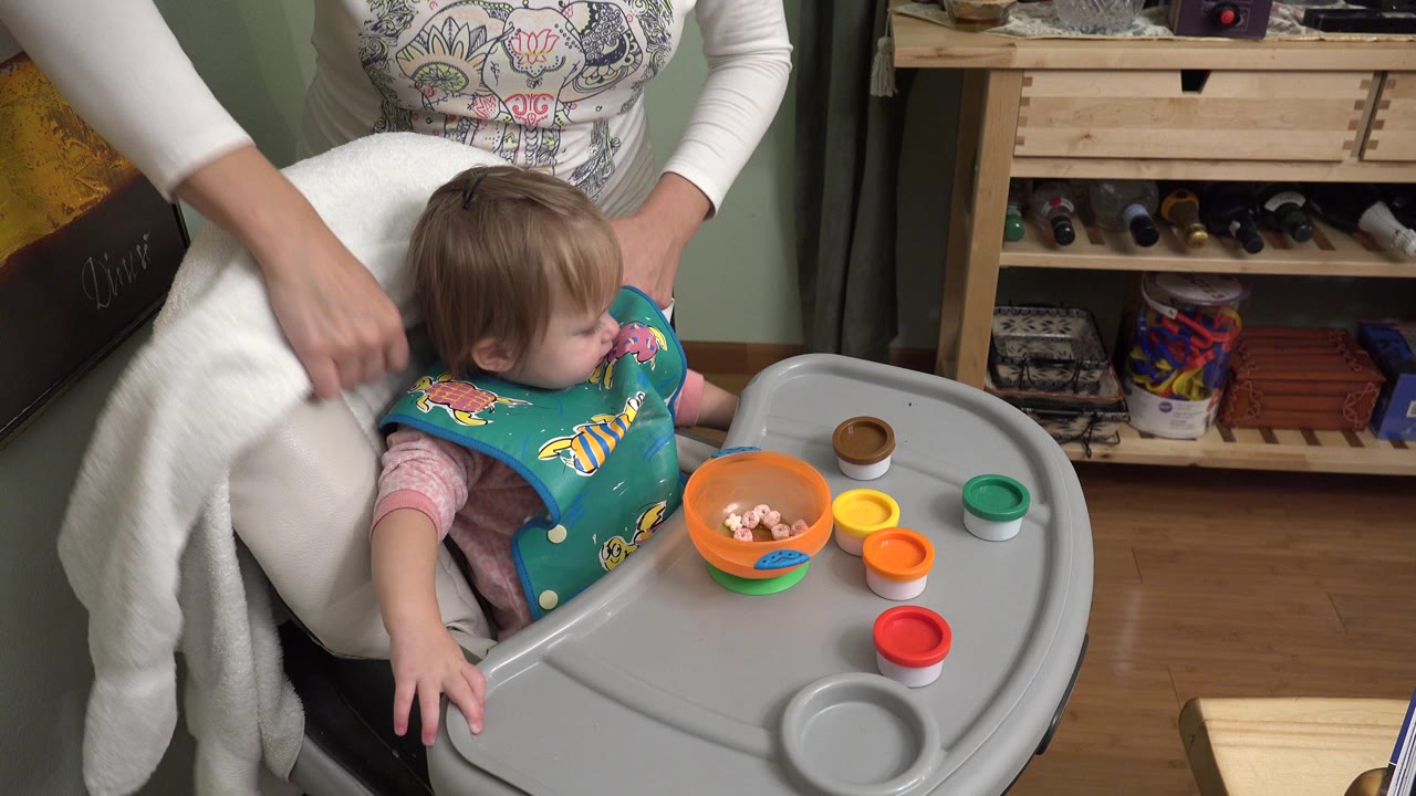 The baby turns left in the high chair while an adult woman's hand rests near the chair and small colored containers sit on the tray in the Westminster home interior.