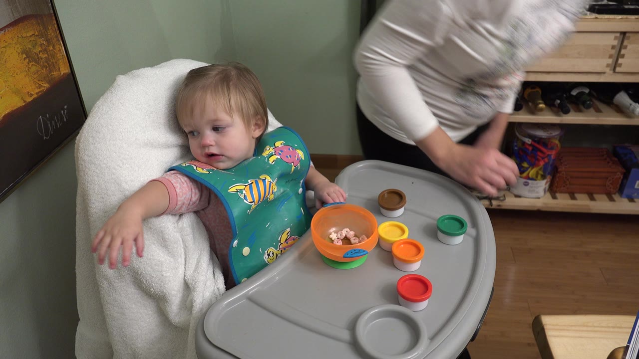 In Westminster, Colorado, the baby extends one arm from the high chair as an adult bends over the tray area with food and colored lids visible.