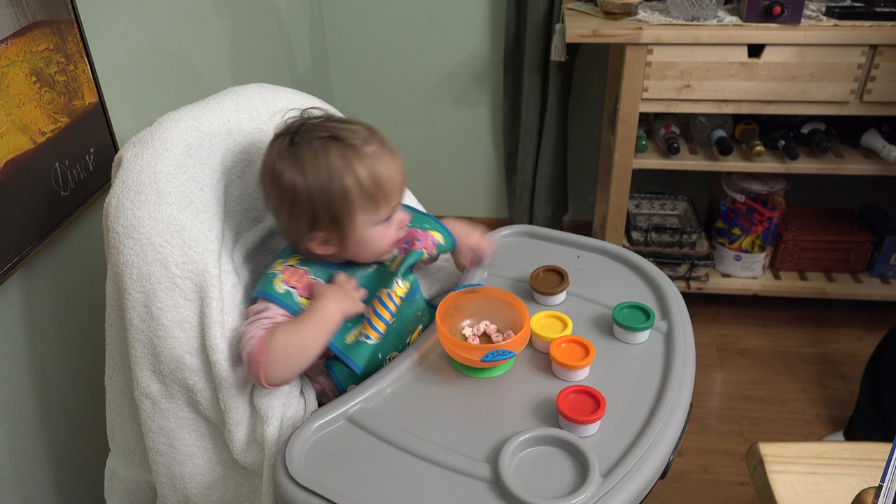 At 0:01 in Westminster, Colorado, the baby turns slightly in the high chair while the tray holds an orange bowl, small round lids, and a cup area.