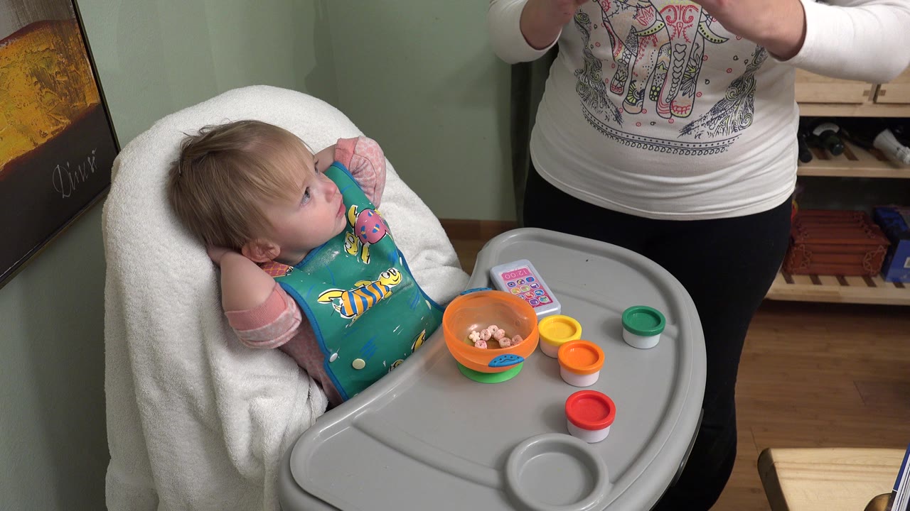 In Westminster, Colorado, a toddler sits in a high chair with a teal bib while an adult stands beside red, orange, yellow, and green play dough containers on the tray.
