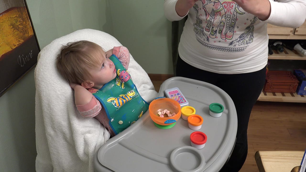 At 0:01 in the Westminster home interior, the toddler looks up from the high chair as an adult's hands appear above the tray with colorful play dough containers nearby.