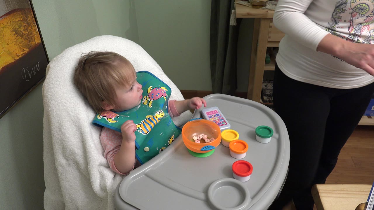 A baby in Westminster, Colorado sits in a high chair with a teal bib, an orange bowl, colorful toy cups, and an adult partly visible at the right edge.