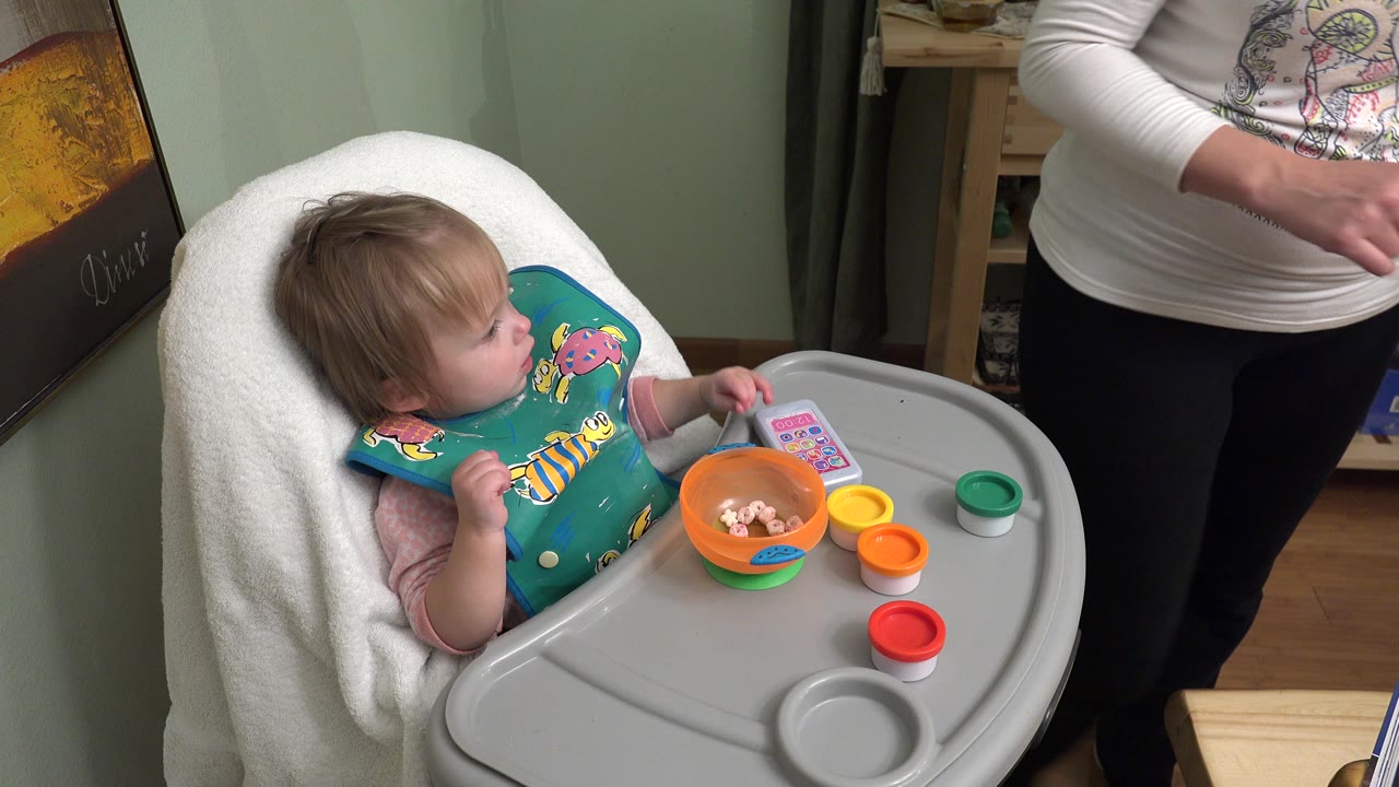 A baby in a high chair in Westminster, Colorado looks toward a partially visible adult, with an orange bowl and red, orange, yellow, and green toy cups on the tray.