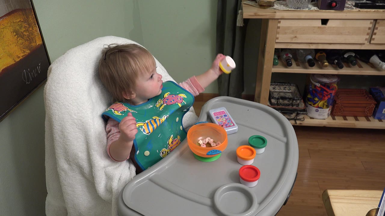 Toddler in a high chair holding a small cup at a tray.