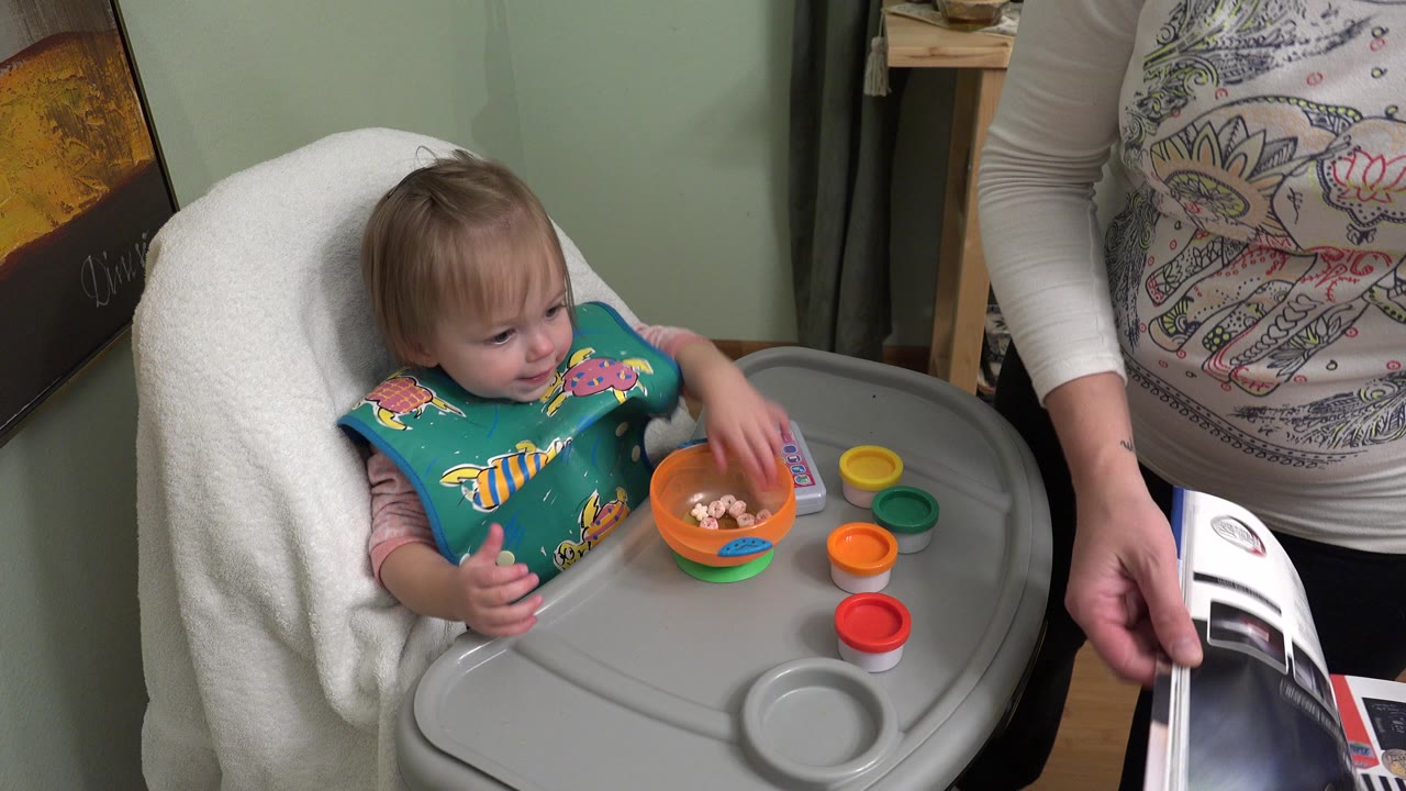 In Westminster, Colorado, a baby sits in a high chair with a teal bib, an orange bowl on the tray, and a cropped adult holding printed pages at the right edge.
