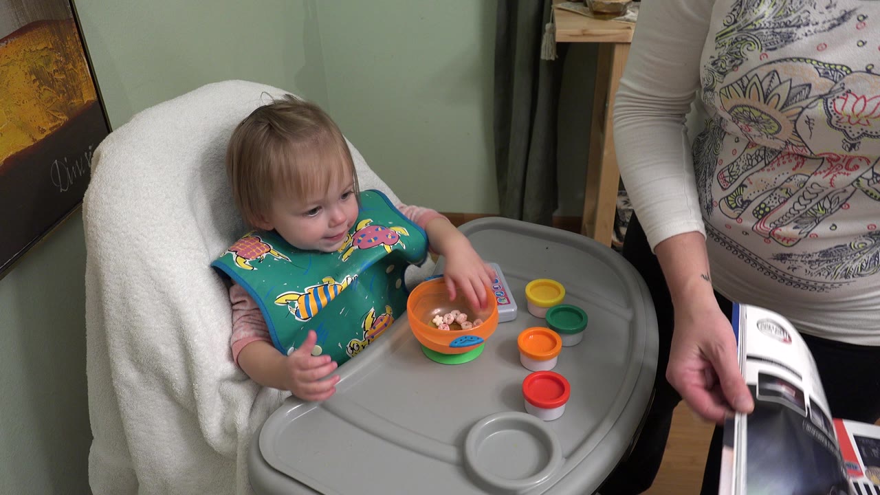 In Westminster, Colorado, the baby looks down while reaching toward food in an orange bowl on the gray high chair tray, with colored cups lined up nearby.