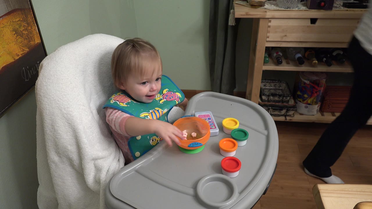 A blond baby in Westminster, Colorado sits in a high chair and reaches toward an orange bowl while colorful cups and a toy phone sit on the tray.