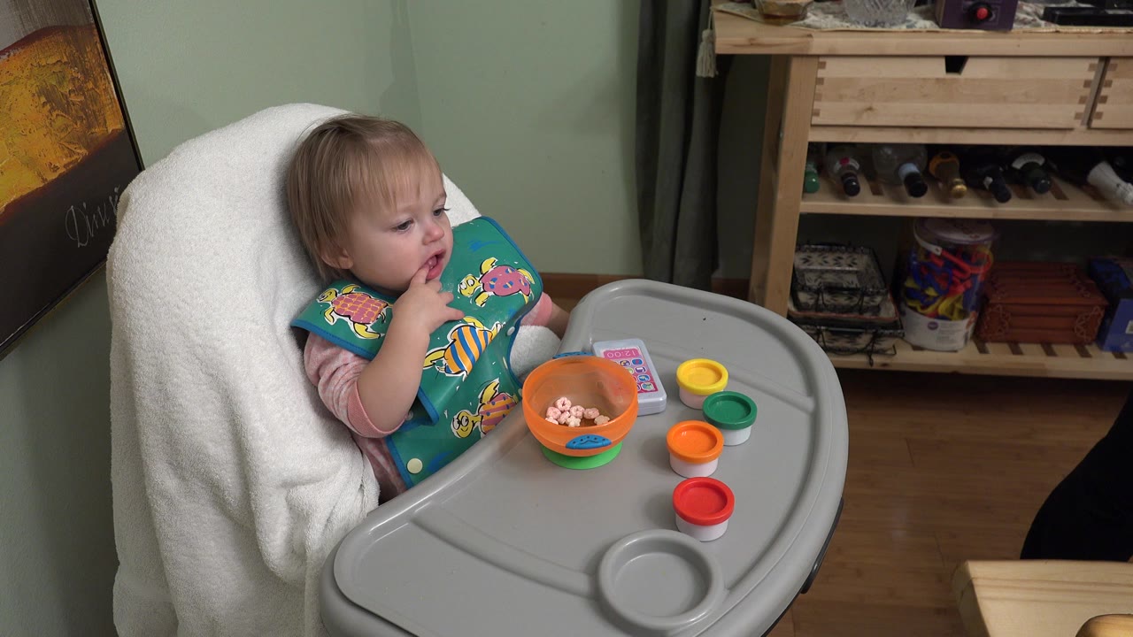 Baby girl in a high chair with a bowl and colored lids on the tray.