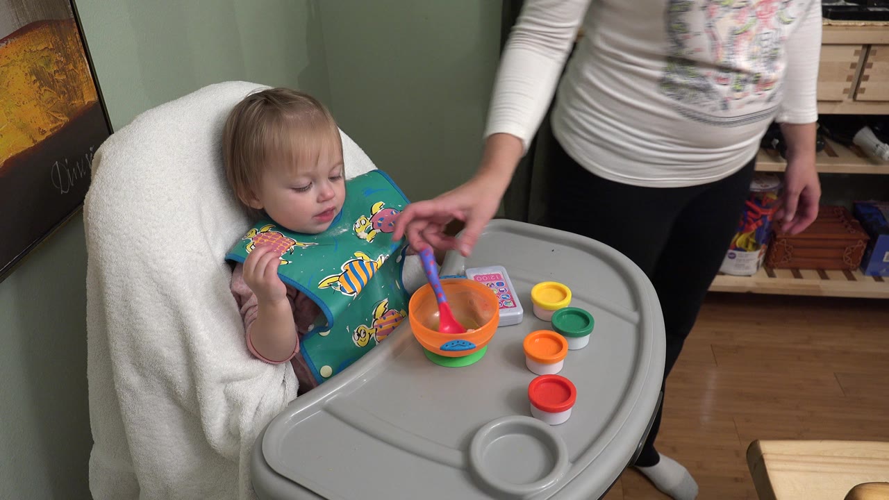 Toddler in a high chair holding utensils beside an adult.