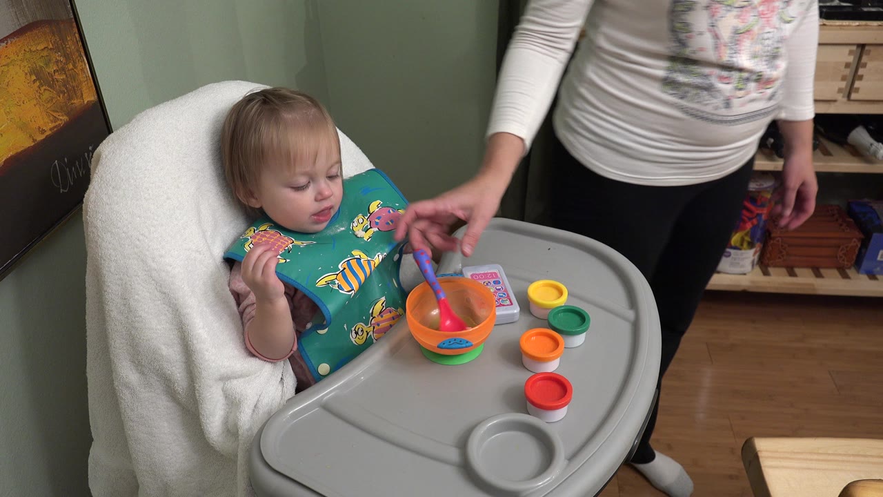 In Westminster, Colorado, the child looks down at the high chair tray as an adult points near the spoon and orange bowl, with small colored lids arranged to the side.
